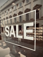 A storefront window displays the word 'SALE' in large white letters. The reflection on the glass shows a street scene with an old, classical-style building, complete with ornate windows. Inside the window, a variety of shoes are organized in a neat display.