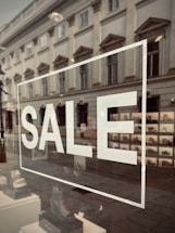 A storefront window displays the word 'SALE' in large white letters. The reflection on the glass shows a street scene with an old, classical-style building, complete with ornate windows. Inside the window, a variety of shoes are organized in a neat display.
