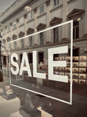 A storefront window displays the word 'SALE' in large white letters. The reflection on the glass shows a street scene with an old, classical-style building, complete with ornate windows. Inside the window, a variety of shoes are organized in a neat display.