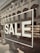 A storefront window displays the word 'SALE' in large white letters. The reflection on the glass shows a street scene with an old, classical-style building, complete with ornate windows. Inside the window, a variety of shoes are organized in a neat display.