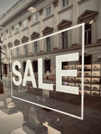 A storefront window displays the word 'SALE' in large white letters. The reflection on the glass shows a street scene with an old, classical-style building, complete with ornate windows. Inside the window, a variety of shoes are organized in a neat display.