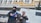 Two individuals dressed in winter attire, including hats and masks, are standing outside a building with Cyrillic lettering. One is holding a blue folder and appears to be engaging in an official capacity. Snow is falling, and there is a van or vehicle in the background.