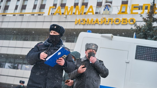 Two individuals dressed in winter attire, including hats and masks, are standing outside a building with Cyrillic lettering. One is holding a blue folder and appears to be engaging in an official capacity. Snow is falling, and there is a van or vehicle in the background.