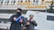 Two individuals dressed in winter attire, including hats and masks, are standing outside a building with Cyrillic lettering. One is holding a blue folder and appears to be engaging in an official capacity. Snow is falling, and there is a van or vehicle in the background.