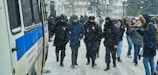 Police officers are escorting a person in a blue jacket with a hood through a snowy street. The officers are wearing dark uniforms with badges and face coverings. Several bystanders, some holding phones or cameras, are present in the background. A large police vehicle is parked nearby.
