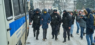 Police officers are escorting a person in a blue jacket with a hood through a snowy street. The officers are wearing dark uniforms with badges and face coverings. Several bystanders, some holding phones or cameras, are present in the background. A large police vehicle is parked nearby.