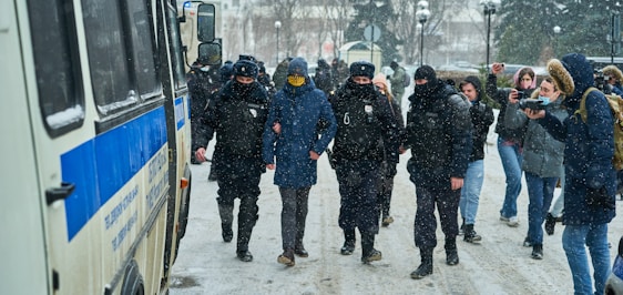 Police officers are escorting a person in a blue jacket with a hood through a snowy street. The officers are wearing dark uniforms with badges and face coverings. Several bystanders, some holding phones or cameras, are present in the background. A large police vehicle is parked nearby.
