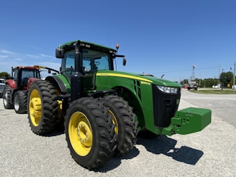 A large green tractor with yellow wheels is parked on a gravel surface. It features the model number 8360 R clearly visible and is situated outdoors under a clear blue sky. Another red tractor is parked beside it, and trees and streetlights are visible in the background.