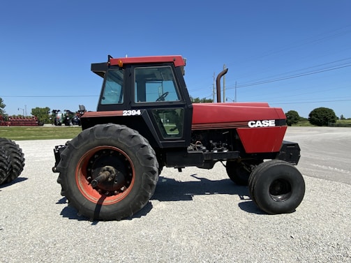 A red and black tractor is parked on a gravel surface under a clear blue sky. The tractor has large black tires, a cab with windows, and displays the brand name 'CASE' along with the model number '2394' on the side. In the background, there are more agricultural machines and some greenery.