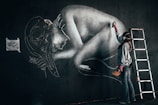 A street artist is painting a large mural on a dark wall, depicting a curled-up human figure. The artist is wearing protective gear including a mask and gloves, and is using a spray can. There is a metal ladder beside the artist, indicating that they may have been working on higher sections of the wall. A small reference image of the artwork is taped to the wall.