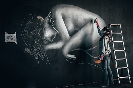 A street artist is painting a large mural on a dark wall, depicting a curled-up human figure. The artist is wearing protective gear including a mask and gloves, and is using a spray can. There is a metal ladder beside the artist, indicating that they may have been working on higher sections of the wall. A small reference image of the artwork is taped to the wall.
