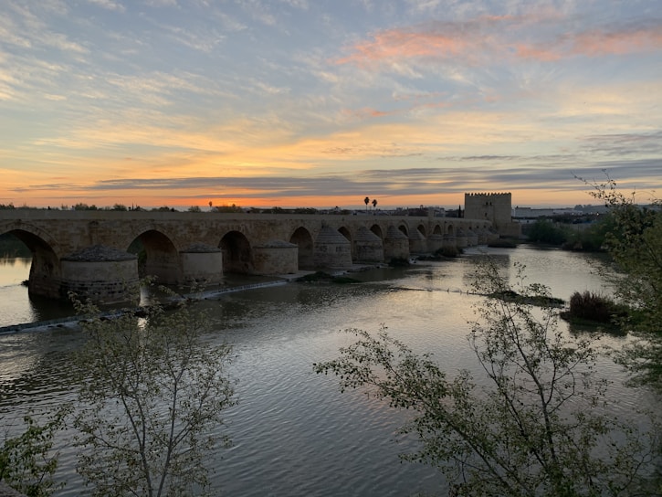 A serene ancient stone bridge arching over a calm river at sunset.