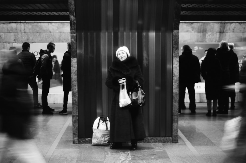 Photo en niveaux de gris d’une femme en manteau noir debout devant des gens dans un immeuble