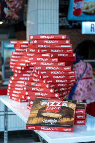A stack of colorful burger and pizza boxes branded with pacaron logo, ready for delivery in a bustling kitchen.
