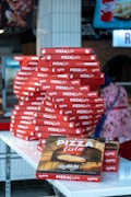 A large stack of red pizza boxes is neatly arranged on a white countertop. The boxes are branded with 'Pizza Lista' and feature designs including a pizza oven. A figure in the background is partially visible, wearing colorful attire, adding to the bustling atmosphere.