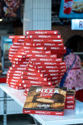 A large stack of red pizza boxes is neatly arranged on a white countertop. The boxes are branded with 'Pizza Lista' and feature designs including a pizza oven. A figure in the background is partially visible, wearing colorful attire, adding to the bustling atmosphere.