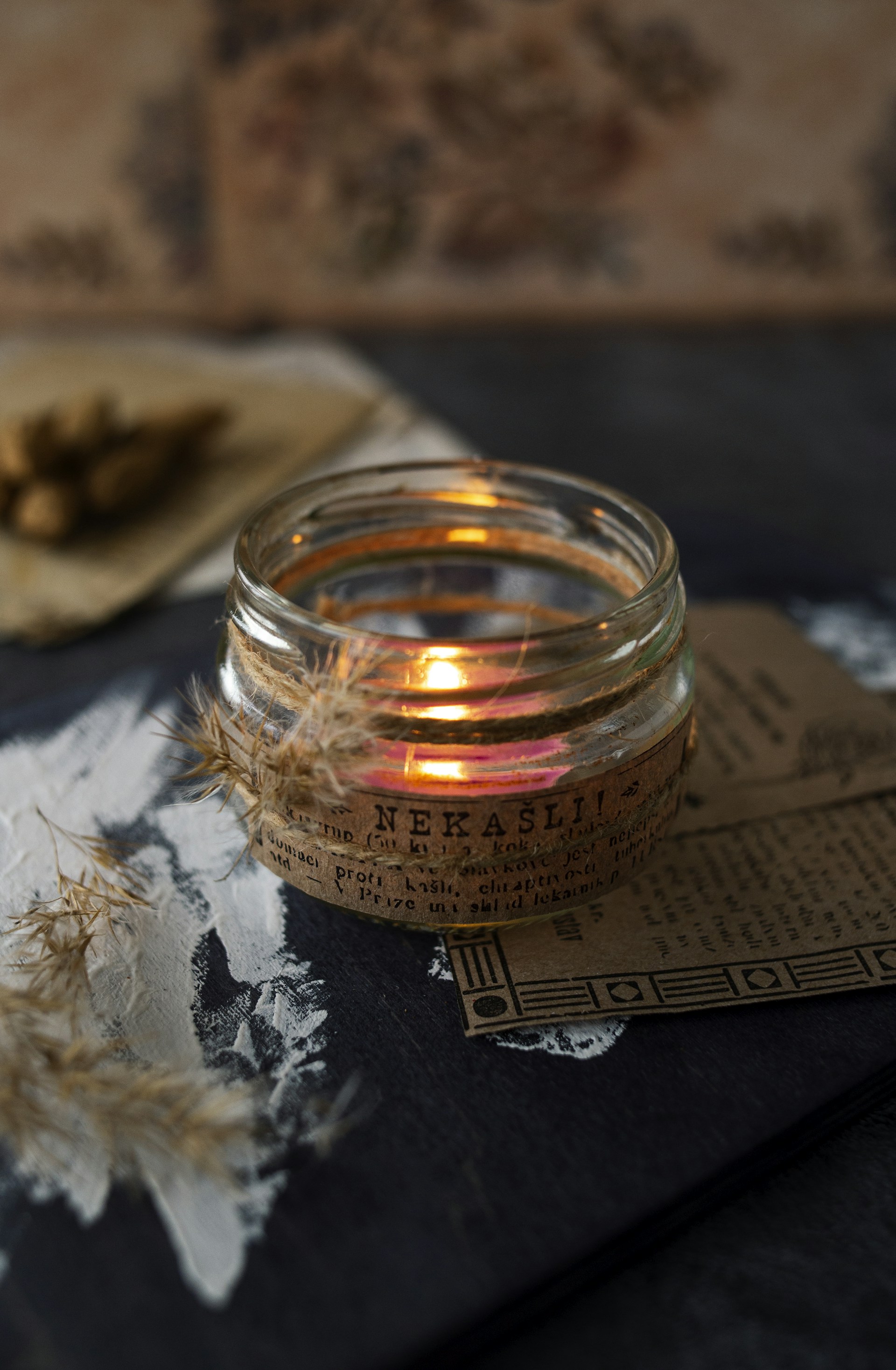 A close-up of a sleek, frosted glass jar candle glowing softly on a wooden table, surrounded by delicate dried flowers.