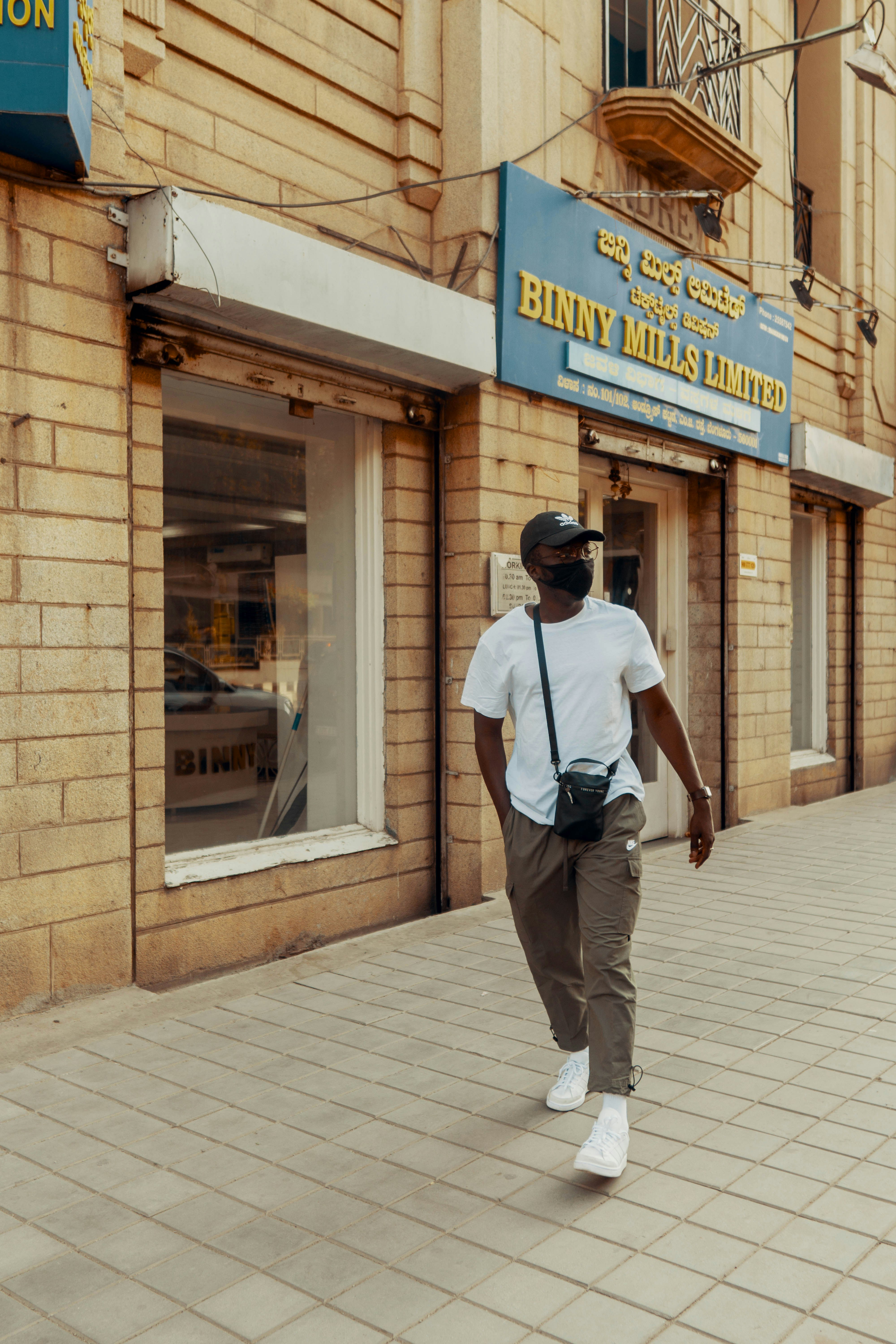 man in white t-shirt and brown pants wearing black backpack walking on sidewalk during daytime