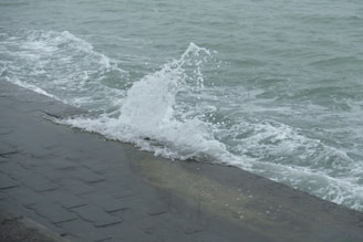 Waves are crashing against a stone seawall, with water splashing upward. The ocean appears choppy with visible whitecaps, and the surface of the water is a muted grey-green.