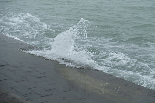 Waves are crashing against a stone seawall, with water splashing upward. The ocean appears choppy with visible whitecaps, and the surface of the water is a muted grey-green.
