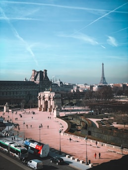 A bustling cityscape with the Eiffel Tower in the distance. The foreground features a busy street with vehicles including buses and cars. A historical arch stands prominently, surrounded by pedestrians walking on a pinkish square. Leafless trees border the open space, suggesting a season other than summer. The sky is clear blue with visible contrails.