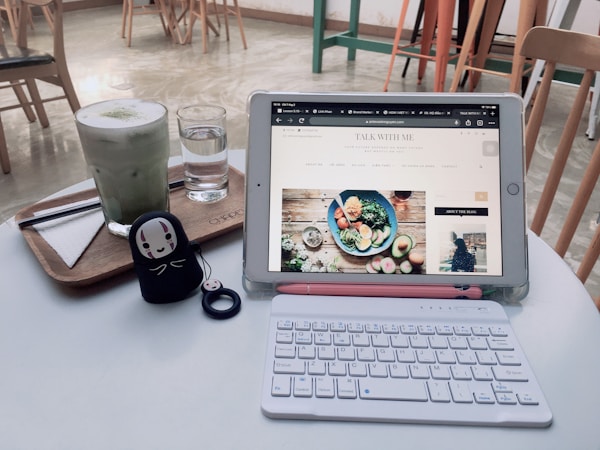 A cozy cafe setting featuring a tablet displaying a food blog, resting on a white table alongside a small, white wireless keyboard. On the table, there is also a wooden tray with a matcha latte and a glass of water. A cute figurine with a face design stands near the keyboard. Wooden chairs and a polished floor are visible in the background, giving a warm and inviting atmosphere.