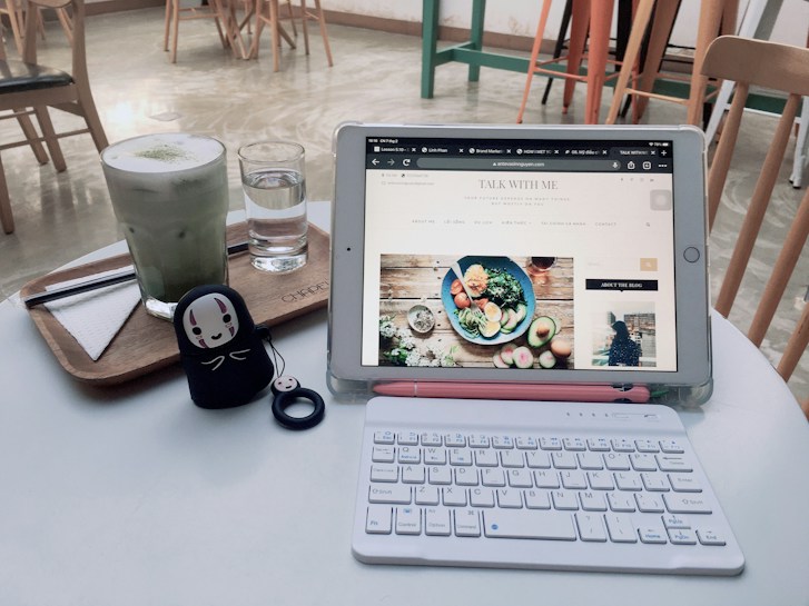 A cozy cafe setting featuring a tablet displaying a food blog, resting on a white table alongside a small, white wireless keyboard. On the table, there is also a wooden tray with a matcha latte and a glass of water. A cute figurine with a face design stands near the keyboard. Wooden chairs and a polished floor are visible in the background, giving a warm and inviting atmosphere.
