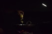 A hand is pouring oil from a bottle into a dark pan or wok on a stovetop. The scene is dimly lit with a focused light beam illuminating the area. The light source is positioned above, creating a moody and intimate atmosphere.