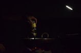 A hand is pouring oil from a bottle into a dark pan or wok on a stovetop. The scene is dimly lit with a focused light beam illuminating the area. The light source is positioned above, creating a moody and intimate atmosphere.