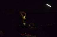 A hand is pouring oil from a bottle into a dark pan or wok on a stovetop. The scene is dimly lit with a focused light beam illuminating the area. The light source is positioned above, creating a moody and intimate atmosphere.