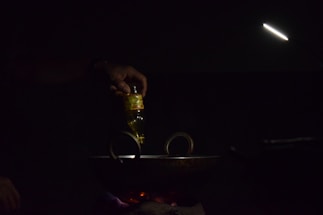 A hand is pouring oil from a bottle into a dark pan or wok on a stovetop. The scene is dimly lit with a focused light beam illuminating the area. The light source is positioned above, creating a moody and intimate atmosphere.
