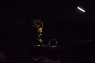 A hand is pouring oil from a bottle into a dark pan or wok on a stovetop. The scene is dimly lit with a focused light beam illuminating the area. The light source is positioned above, creating a moody and intimate atmosphere.