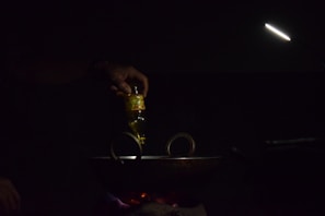 A hand is pouring oil from a bottle into a dark pan or wok on a stovetop. The scene is dimly lit with a focused light beam illuminating the area. The light source is positioned above, creating a moody and intimate atmosphere.