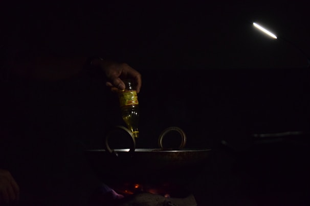 A hand is pouring oil from a bottle into a dark pan or wok on a stovetop. The scene is dimly lit with a focused light beam illuminating the area. The light source is positioned above, creating a moody and intimate atmosphere.