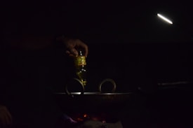 A hand is pouring oil from a bottle into a dark pan or wok on a stovetop. The scene is dimly lit with a focused light beam illuminating the area. The light source is positioned above, creating a moody and intimate atmosphere.