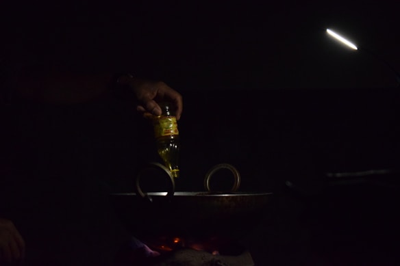A hand is pouring oil from a bottle into a dark pan or wok on a stovetop. The scene is dimly lit with a focused light beam illuminating the area. The light source is positioned above, creating a moody and intimate atmosphere.