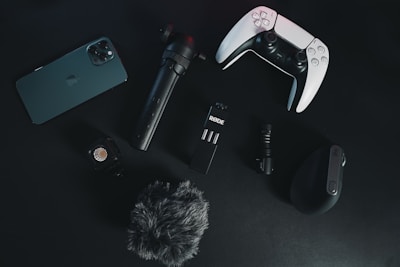 A collection of electronic gadgets is arranged on a dark surface. Items include a smartphone, a video game controller, a microphone, a small tripod, a wireless computer mouse, an action camera, and a furry windscreen cover for a microphone.