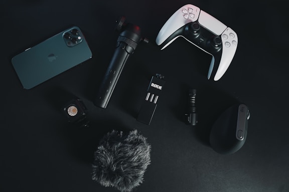 A collection of electronic gadgets is arranged on a dark surface. Items include a smartphone, a video game controller, a microphone, a small tripod, a wireless computer mouse, an action camera, and a furry windscreen cover for a microphone.