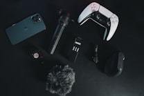 A collection of electronic gadgets is arranged on a dark surface. Items include a smartphone, a video game controller, a microphone, a small tripod, a wireless computer mouse, an action camera, and a furry windscreen cover for a microphone.