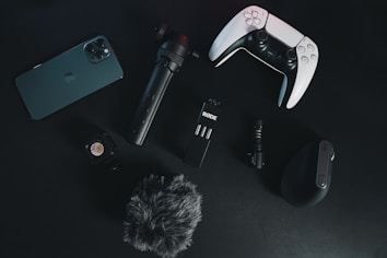 A collection of electronic gadgets is arranged on a dark surface. Items include a smartphone, a video game controller, a microphone, a small tripod, a wireless computer mouse, an action camera, and a furry windscreen cover for a microphone.