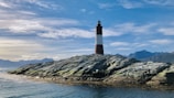 brown and black lighthouse on rocky hill near body of water under blue and white cloudy