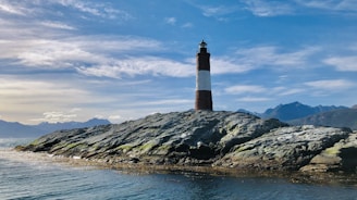brown and black lighthouse on rocky hill near body of water under blue and white cloudy