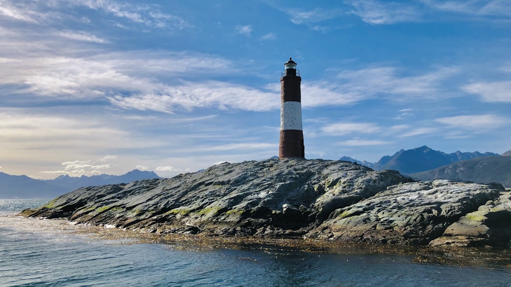 Brauner und schwarzer Leuchtturm auf einem felsigen Hügel in der Nähe eines Gewässers unter blau-weißer Bewölkung