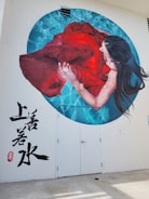 A mural painted on a wall depicts a woman with long dark hair submerged in water, holding a flowing red dress. The water around her is illustrated in shades of blue, creating a calming ripple effect. The mural includes artistic Asian script in black on the white wall to the left.