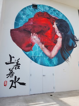 A mural painted on a wall depicts a woman with long dark hair submerged in water, holding a flowing red dress. The water around her is illustrated in shades of blue, creating a calming ripple effect. The mural includes artistic Asian script in black on the white wall to the left.