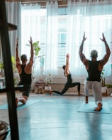 Three people are practicing yoga in a sunlit studio. They are positioned on yoga mats, performing a lunge pose with arms raised towards the ceiling. Large windows with sheer curtains allow natural light to fill the room. Potted plants are placed around the space, adding a touch of greenery to the setting.