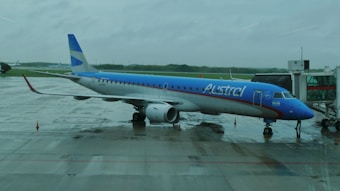 A blue and white commercial airplane is parked at an airport gate on a rainy day. The airplane has the name 'Austral' written on its side. The ground is wet, reflecting the overcast and cloudy weather, and a jet bridge is connected to the airplane for passenger boarding.