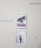 A wall with five pieces of art decor including a sign that says 'Be fearless', a picture of a pink van by palm trees, a sign that says 'Regret Nothing', a picture of an open book with flowers, and a small sign that says 'family forever'. The wall appears to have some tape or marks.