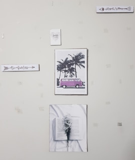 A wall with five pieces of art decor including a sign that says 'Be fearless', a picture of a pink van by palm trees, a sign that says 'Regret Nothing', a picture of an open book with flowers, and a small sign that says 'family forever'. The wall appears to have some tape or marks.