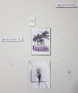A wall with five pieces of art decor including a sign that says 'Be fearless', a picture of a pink van by palm trees, a sign that says 'Regret Nothing', a picture of an open book with flowers, and a small sign that says 'family forever'. The wall appears to have some tape or marks.